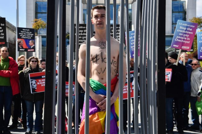 An activist stands naked, wrapped in a rainbow flag, in a mock cage in front of the Chancellery in Berlin on April 30, 2017, during a demonstration calling on Russian President to put an end to the persecution of gay men in Chechnya.  The protestors called on German Chancellor Angela Merkel, who will meet Putin in Sochi on May 2, 2017, to raise the issue with him. / AFP PHOTO / John MACDOUGALL        (Photo credit should read JOHN MACDOUGALL/AFP/Getty Images)